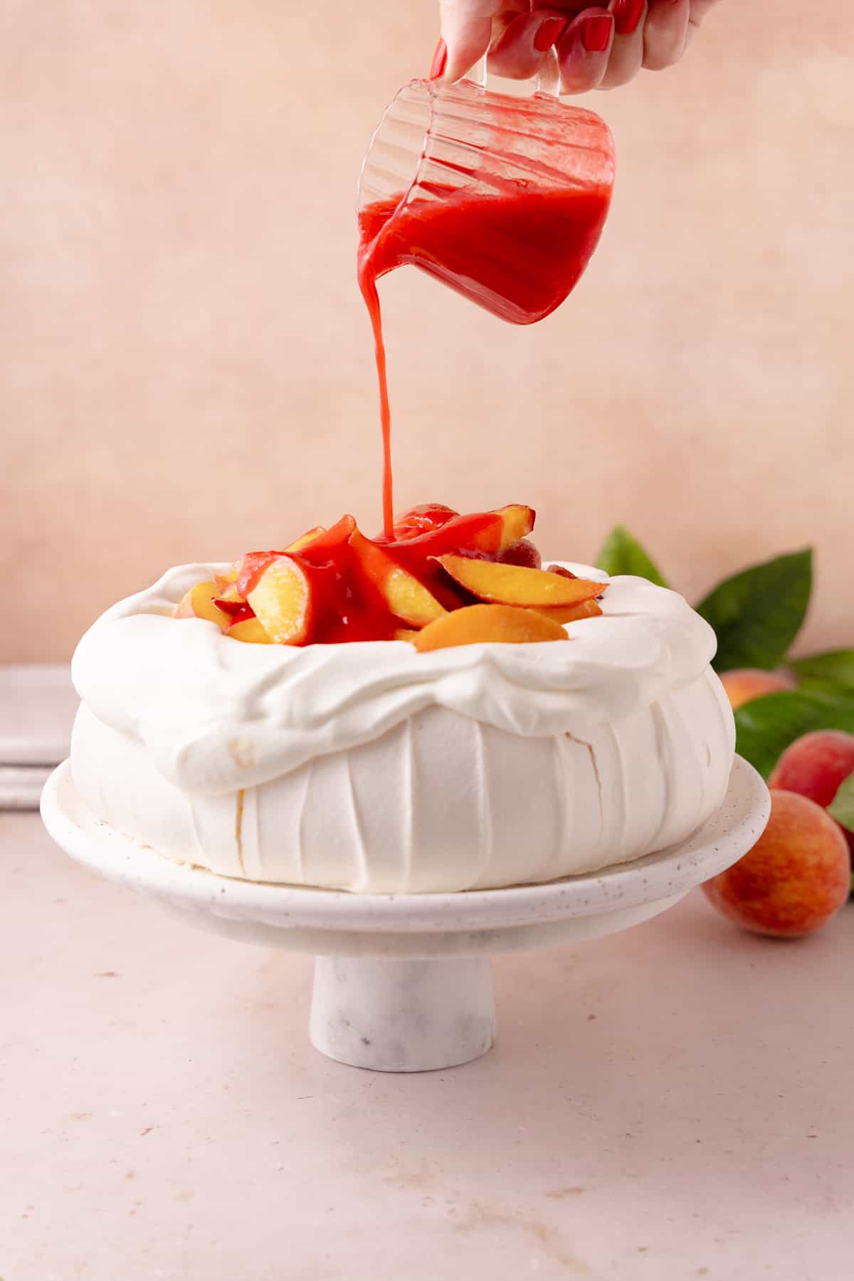 Hand pours raspberry coulis on top of peach melba pavlova.