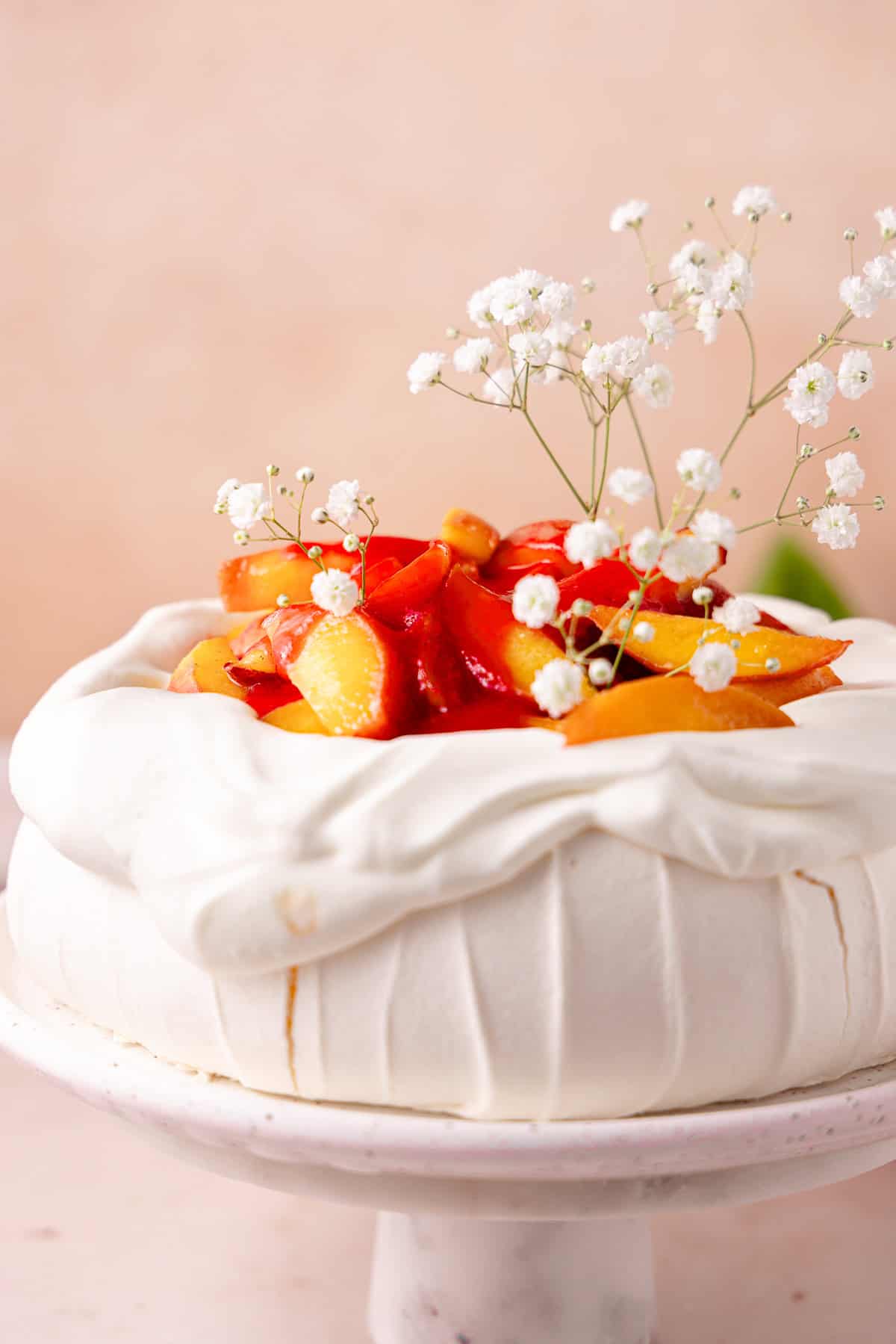 Peach Melba Pavlova topped with whipped cream, peaches and raspberry coulis sitting on a cake stand. 