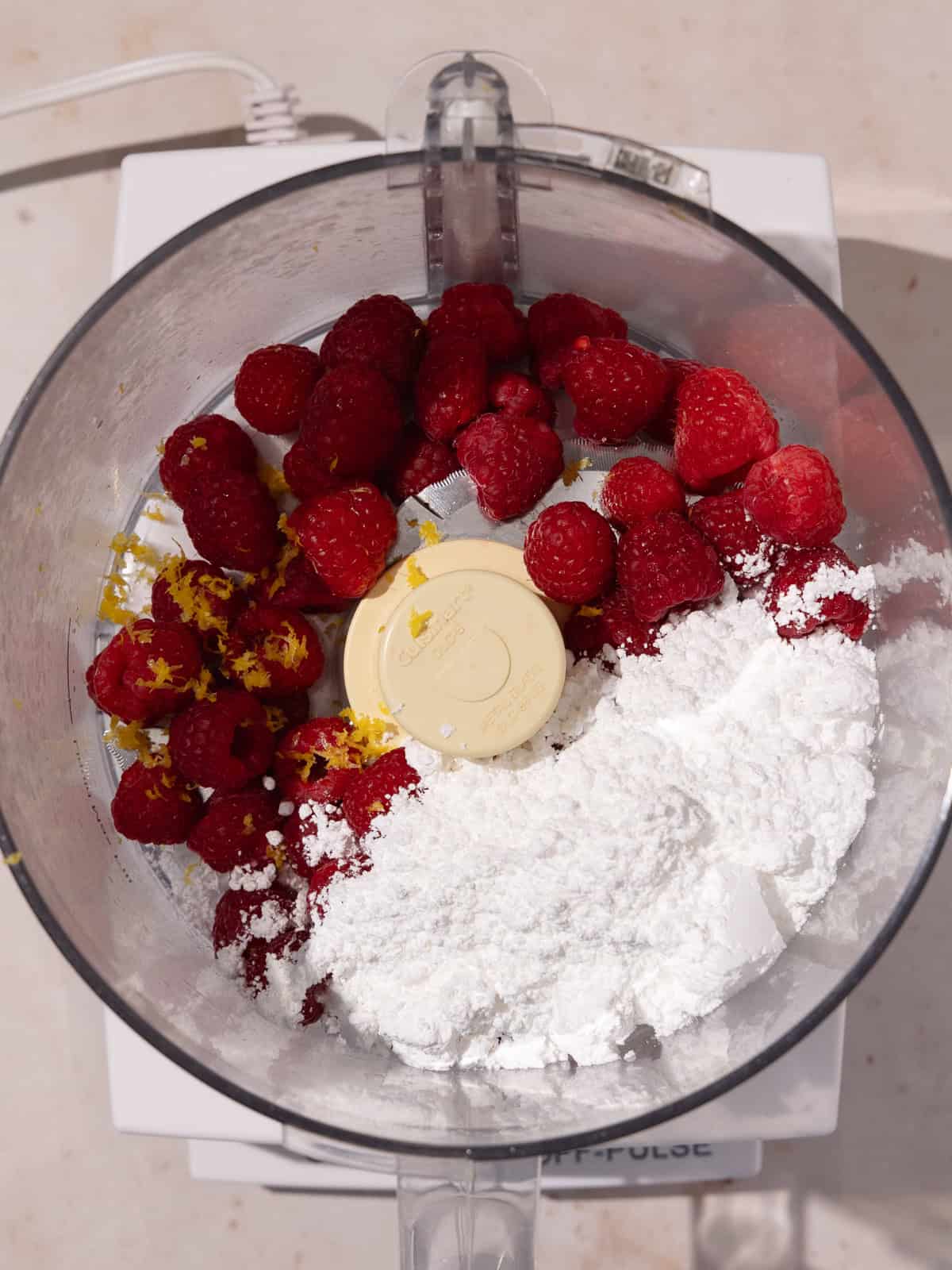 Fresh raspberries, powdered sugar, lemon zest and lemon juice are in the bowl of a food processor.