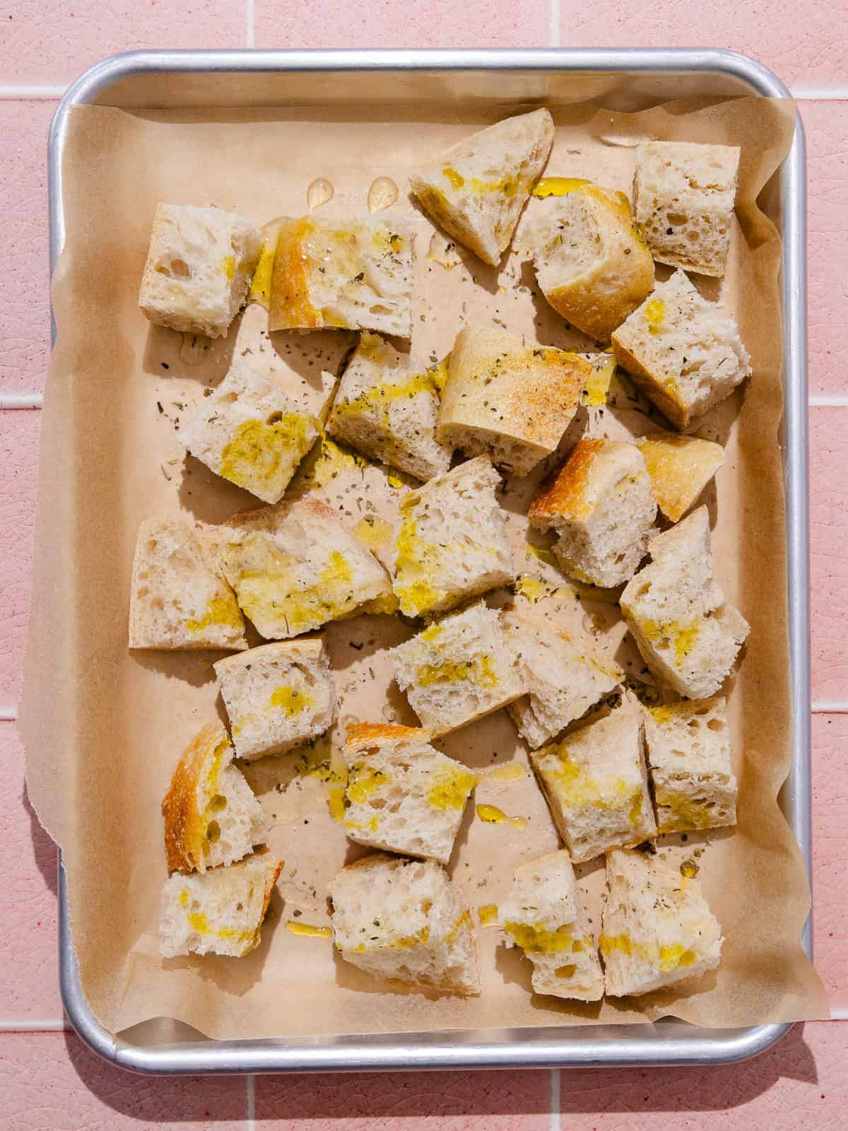 Cubed sourdough bread is tossed with olive oil, italian seasoning and salt on a small baking tray lined with parchment paper.