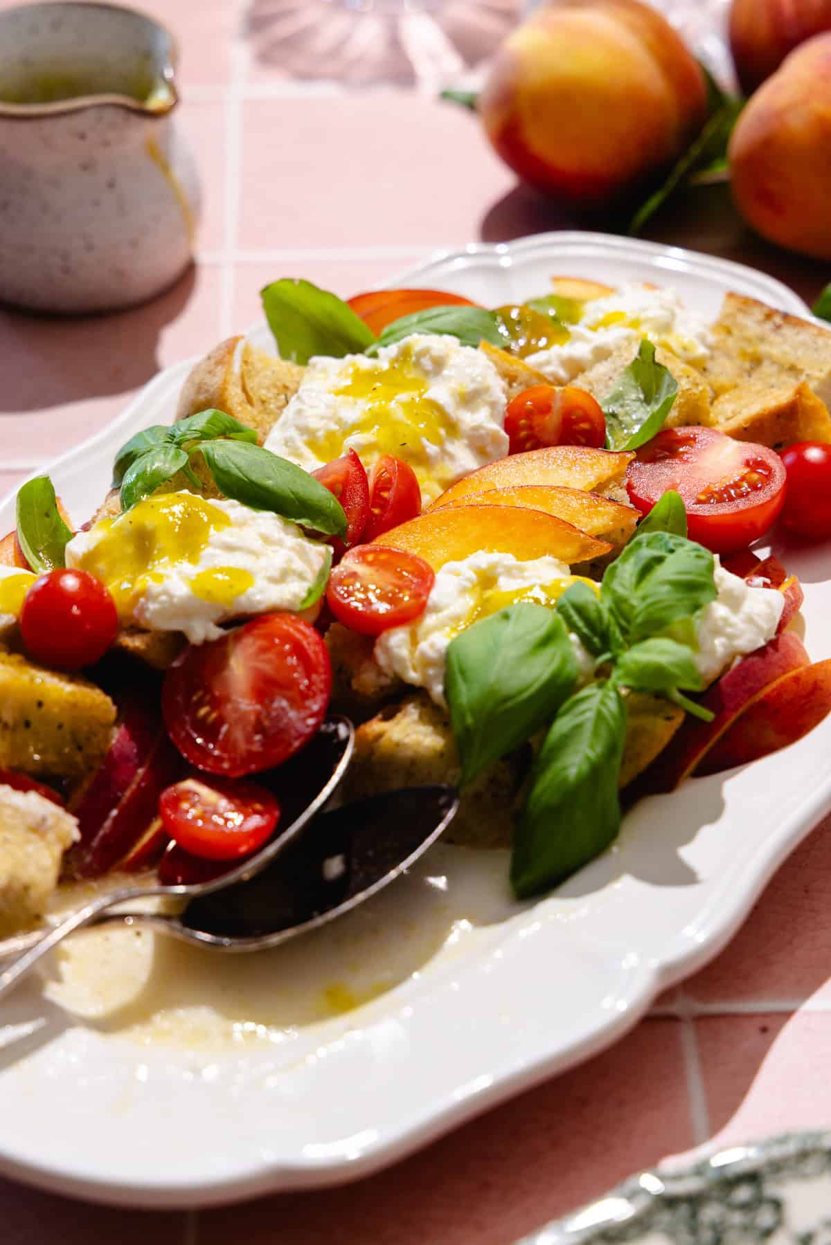 Two serving spoons are on a platter of peach panzanella salad, featuring sliced peaches, cherry tomatoes, homemade croutons, fresh basil and fresh burrata cheese. A creamer filled with vinaigrette is nearby.