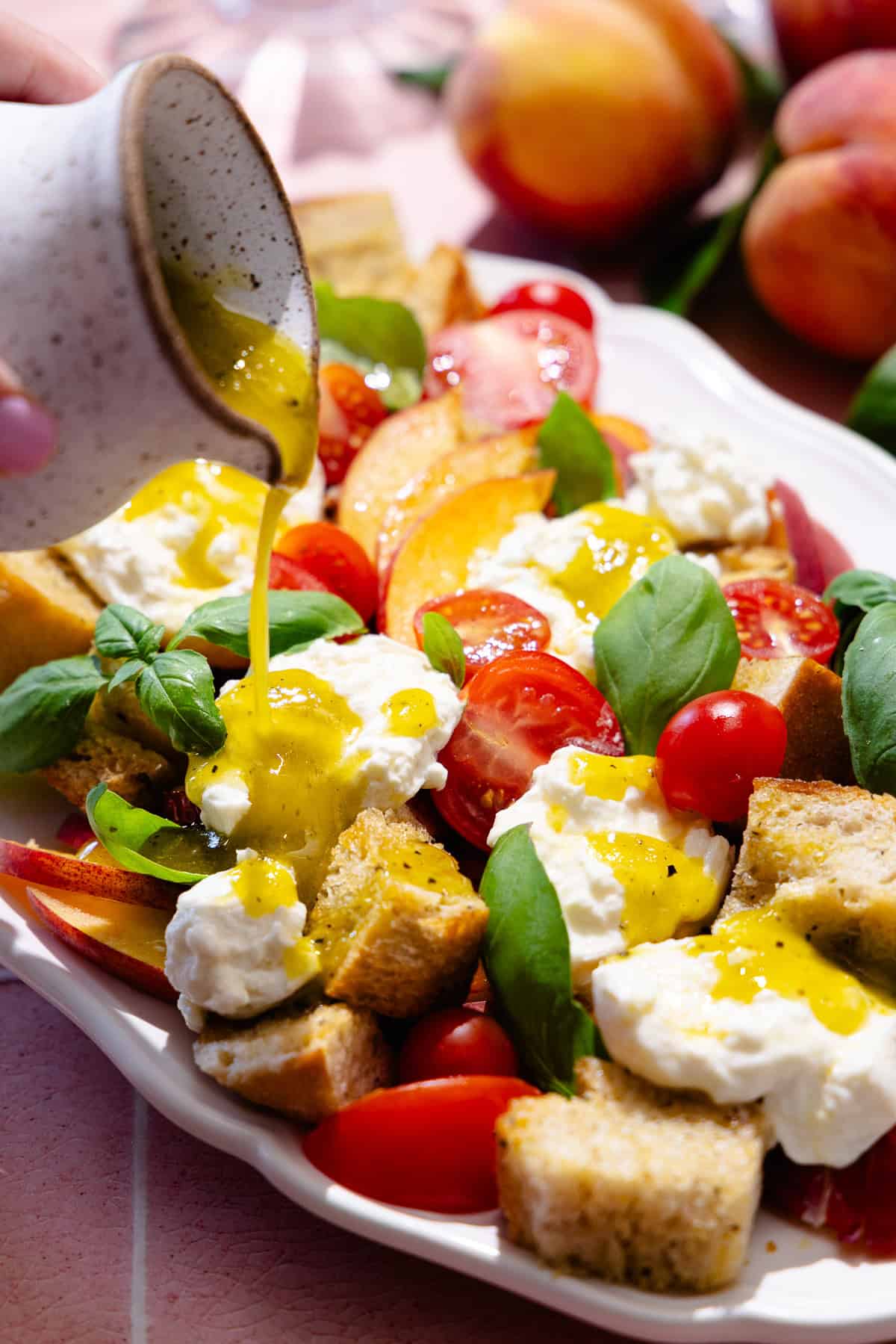 A platter with peach panzanella salad, featuring sliced peaches, cherry tomatoes, homemade croutons, fresh basil and fresh burrata cheese. A hand pours vinaigrette over salad.