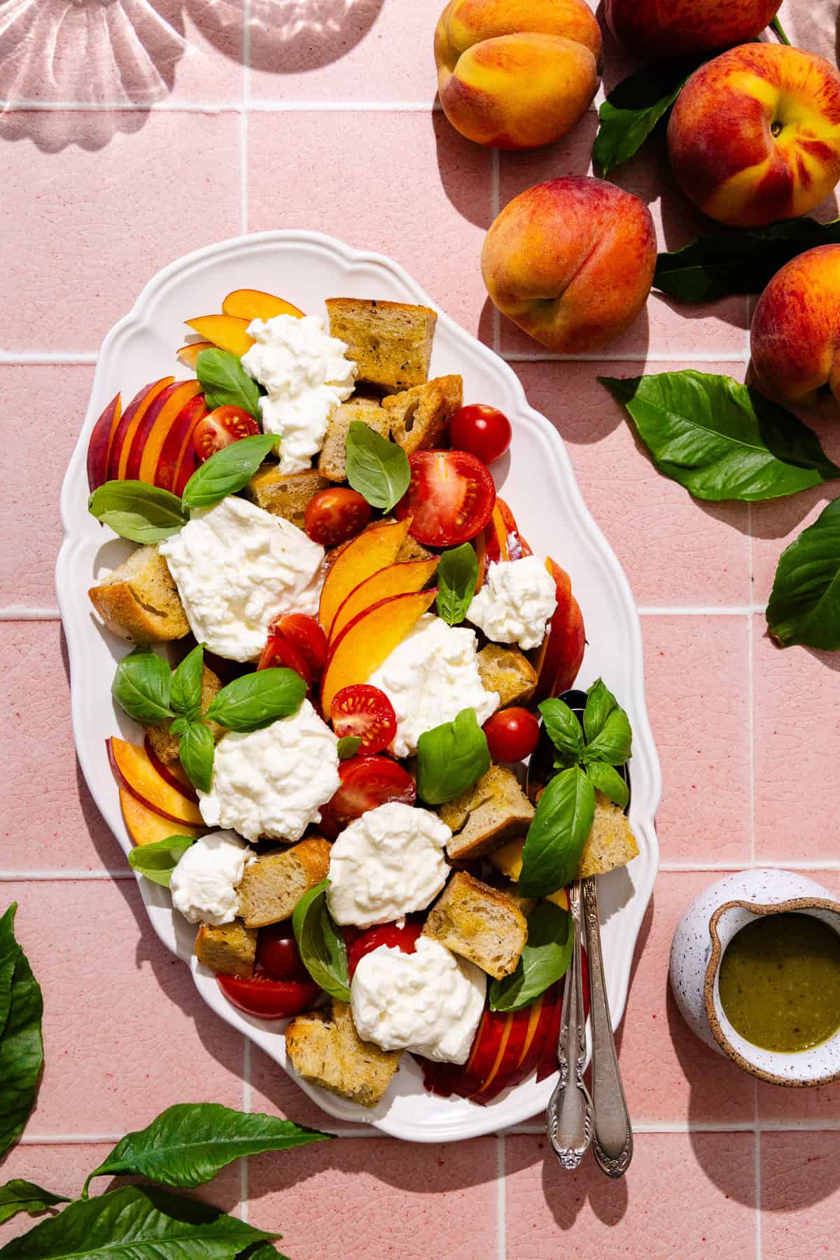 A platter with peach panzanella salad, featuring sliced peaches, cherry tomatoes, homemade croutons, fresh basil and fresh burrata cheese. A creamer filled with vinaigrette is nearby.