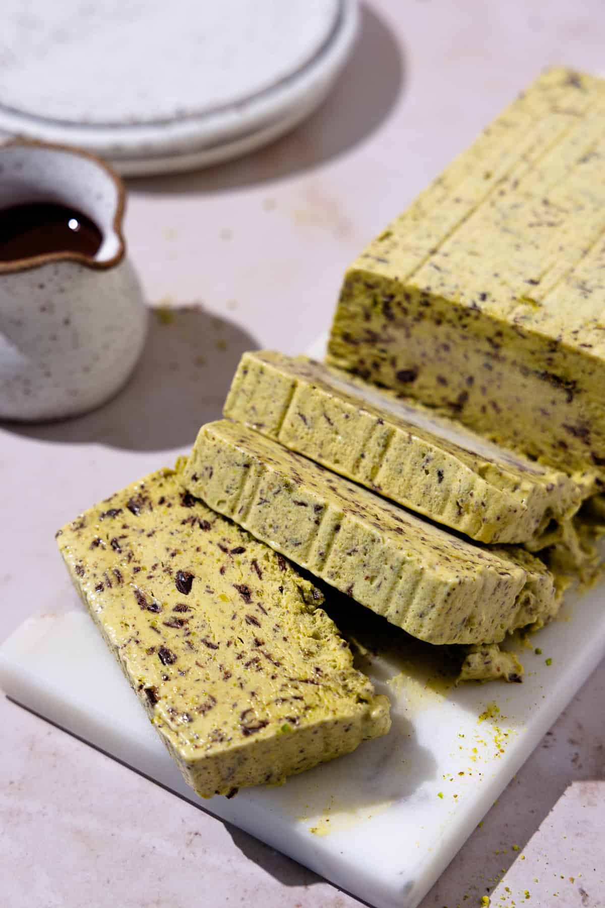 Sliced Pistachio Semifreddo on a marble cutting board. Chocolate sauce in a creamer is nearby.