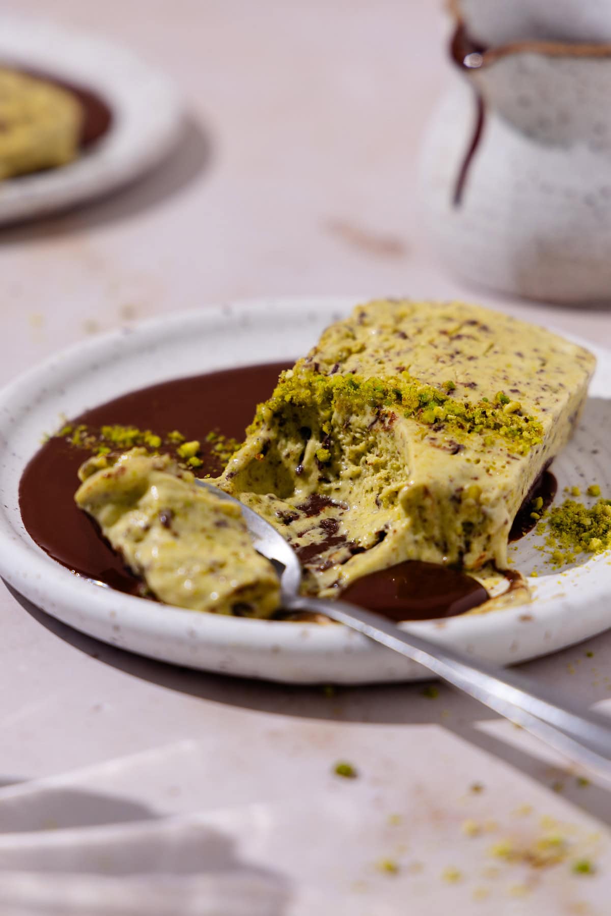 A slice of pistachio semifreddo is on a plate with chocolate sauce. A spoon scoops a bit of semifreddo, showing the creamy texture.