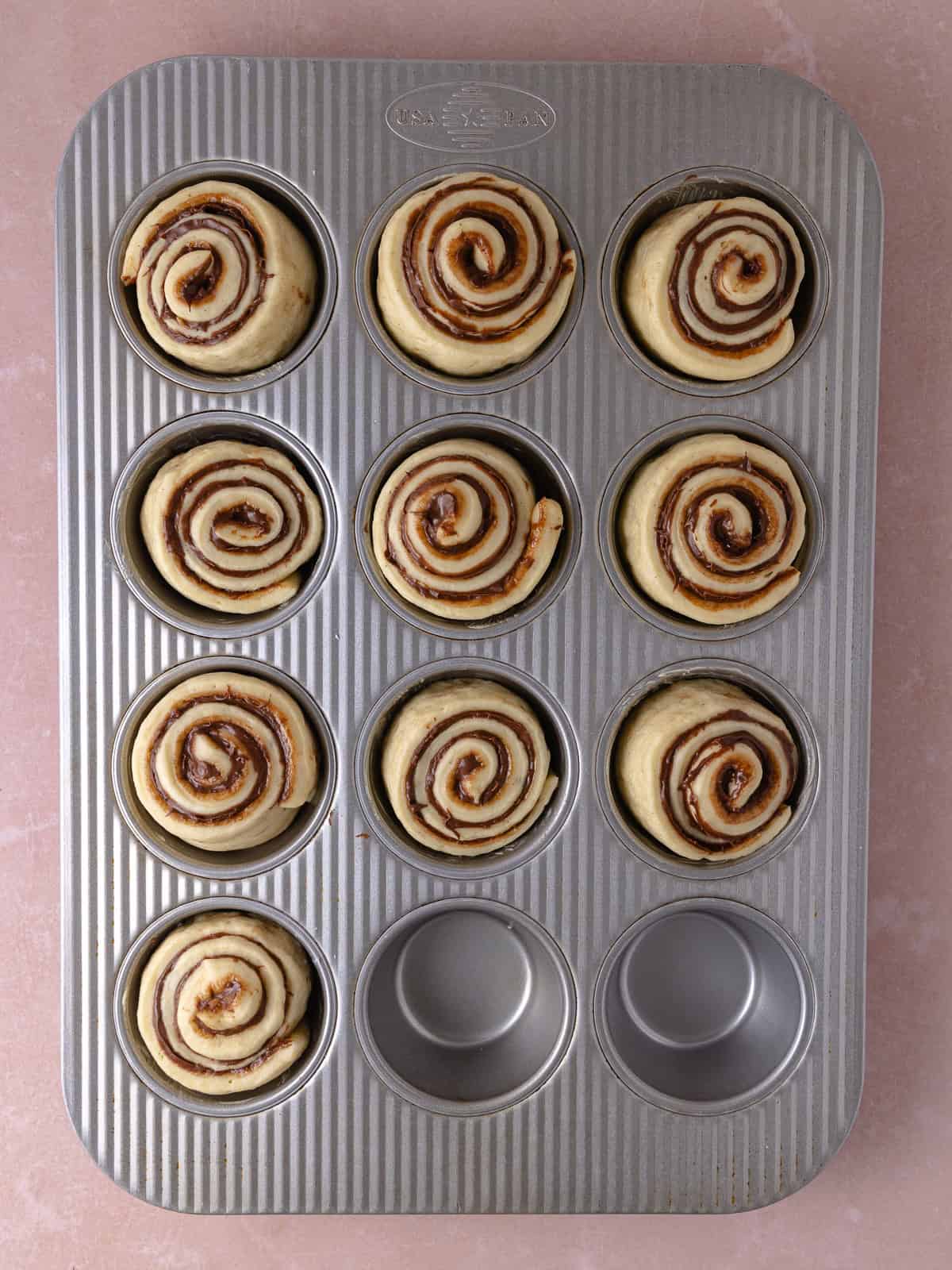 Rolled Nutella buns are placed in a buttered muffin tin.