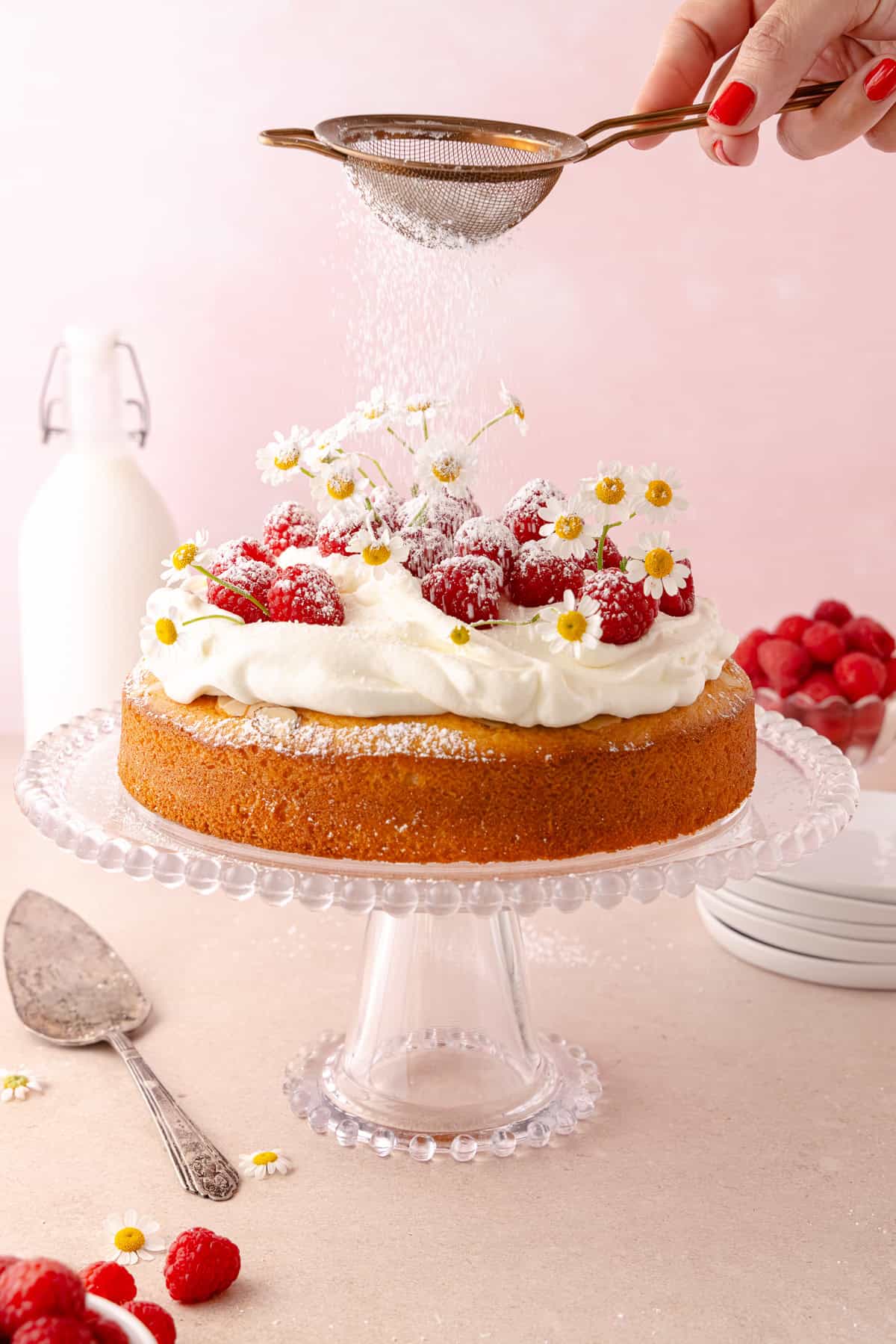 Raspberry olive oil cake sits on a glass cake stand with fresh raspberries all around. A hand with a sifter dusts the cake with powdered sugar.