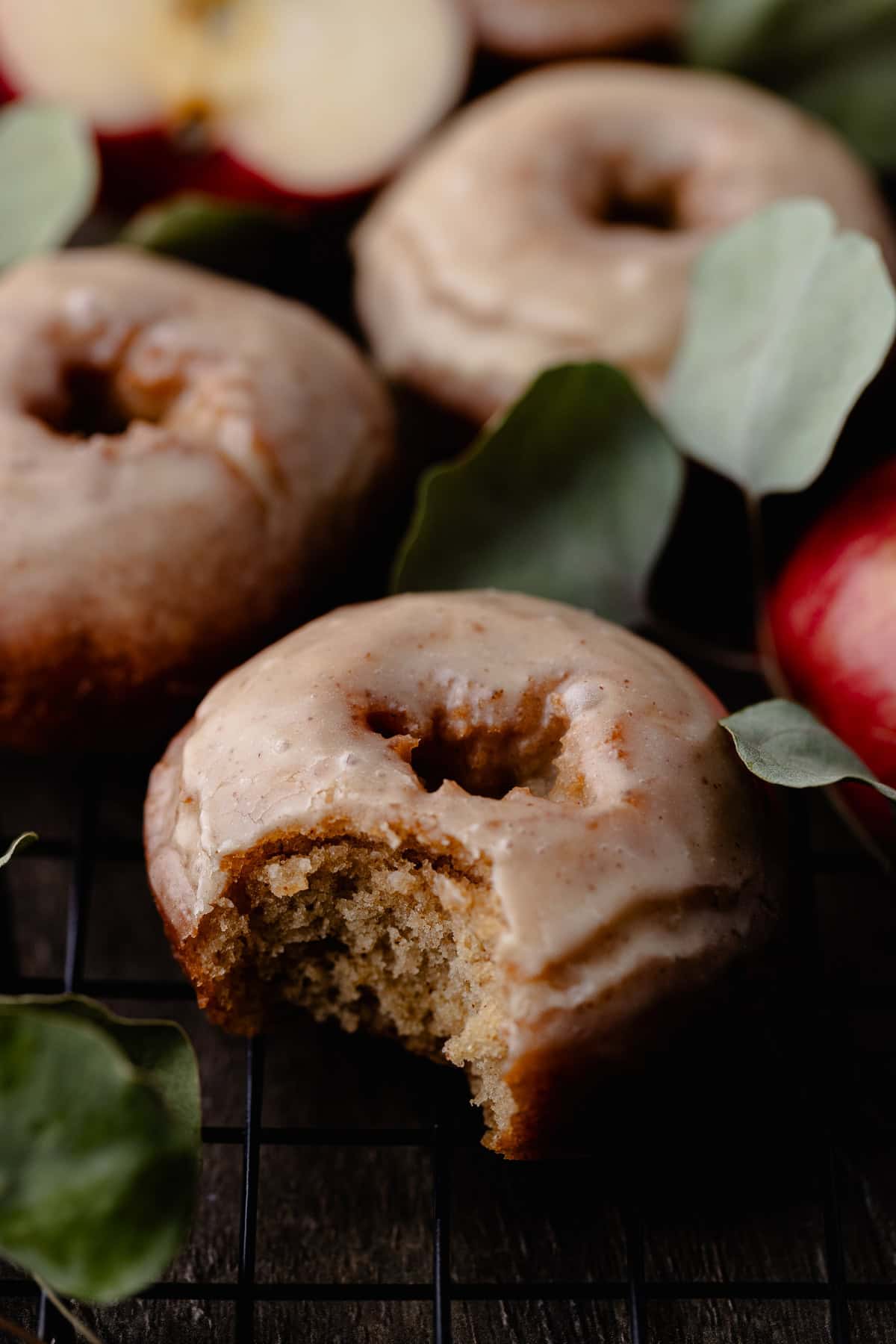 Mini fried apple cider donuts are on a wire rack surrounded by apples. A bite is taken out of a donut.