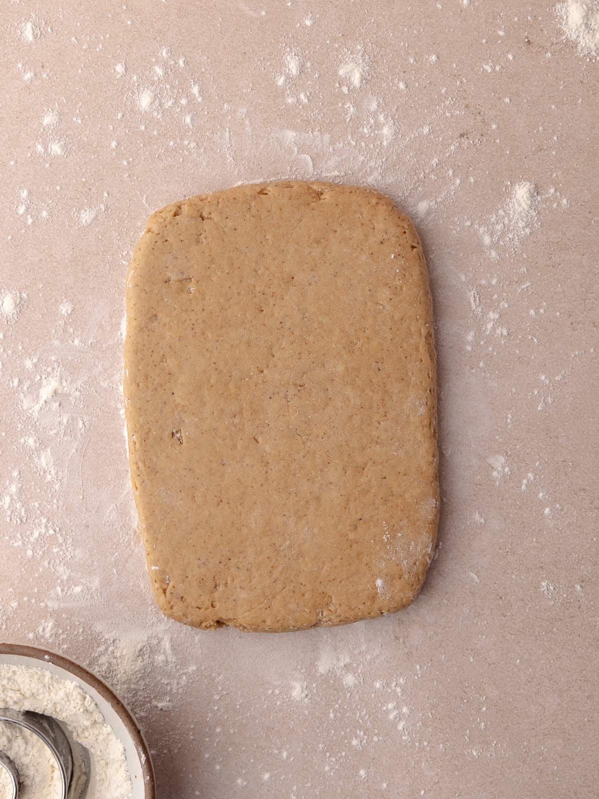 The apple cider donut dough is rolled out to roughly a 6 by 8 inch rectangle. A bowl with flour and biscuit cutters are nearby.