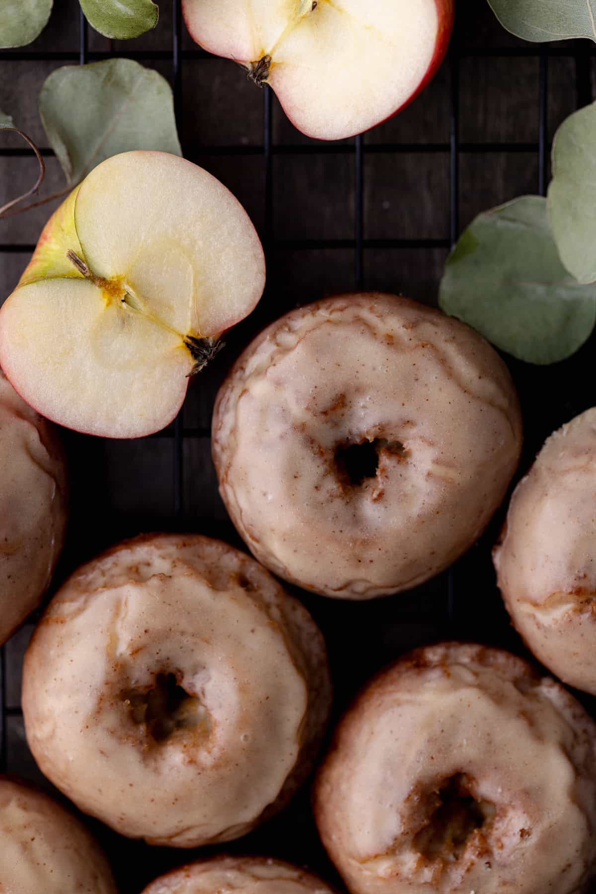 Fried and glazed apple cider donuts are on a wire rack surrounded by apples and dried leaves.