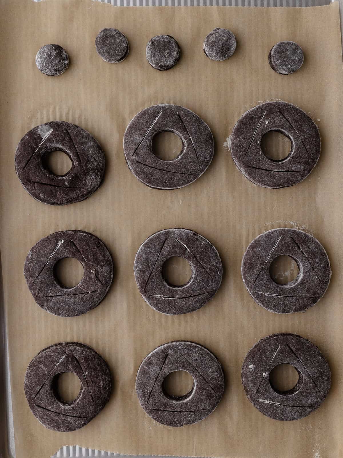 Old Fashioned Chocolate donuts are on a large baking sheet. Each donut is scored with a triangle shape.