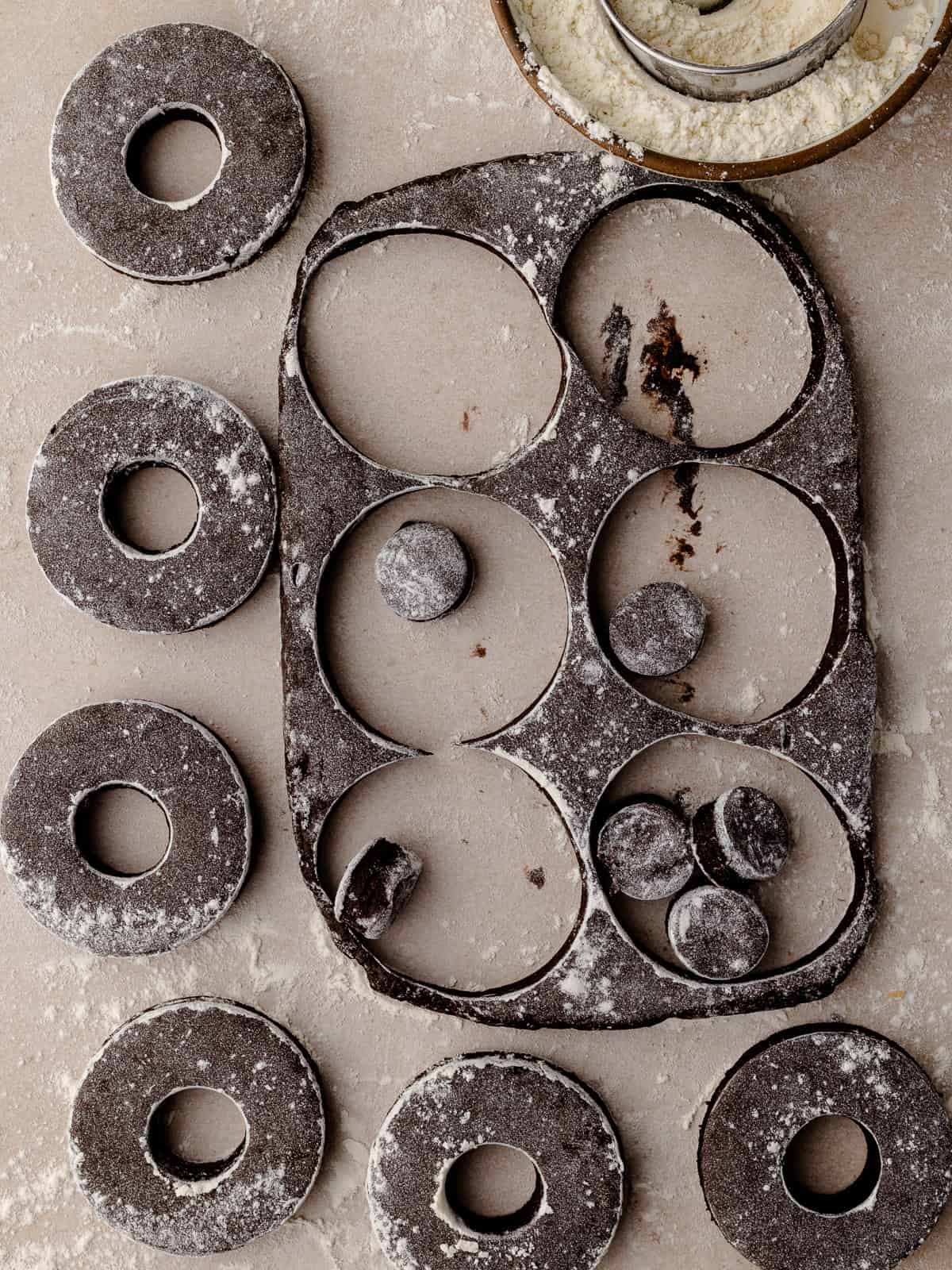 Donuts and donut holes are cut out with round cookie cutters. A bowl of flour is nearby.