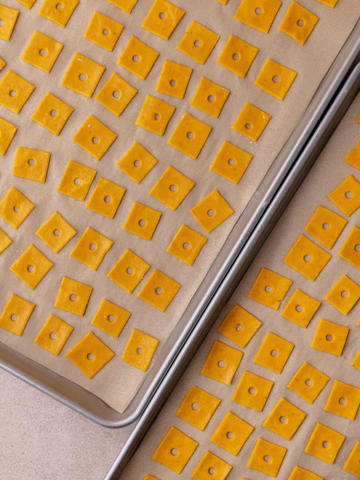 Two baking sheets are filled with unbaked pumpkin cheddar cracker squares and are ready to be baked.