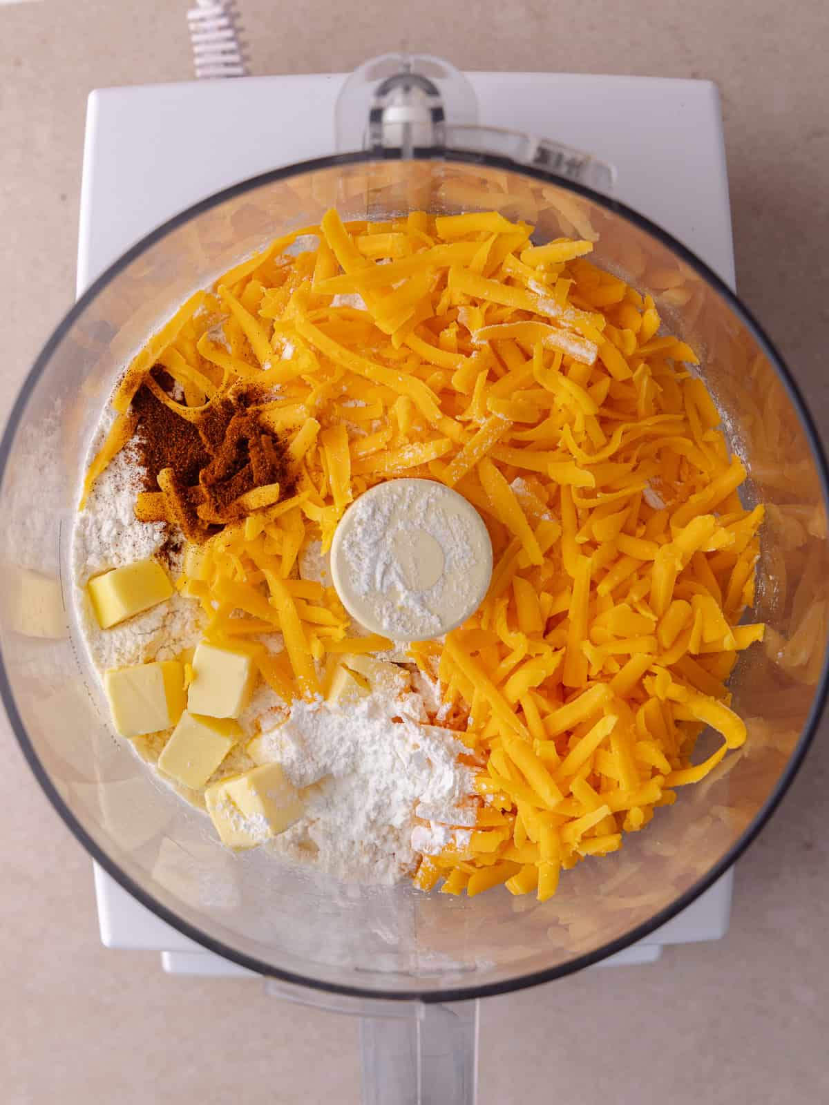 All purpose flour, cornstarch, cheddar cheese, butter and paprika are in the bowl of a food processor.