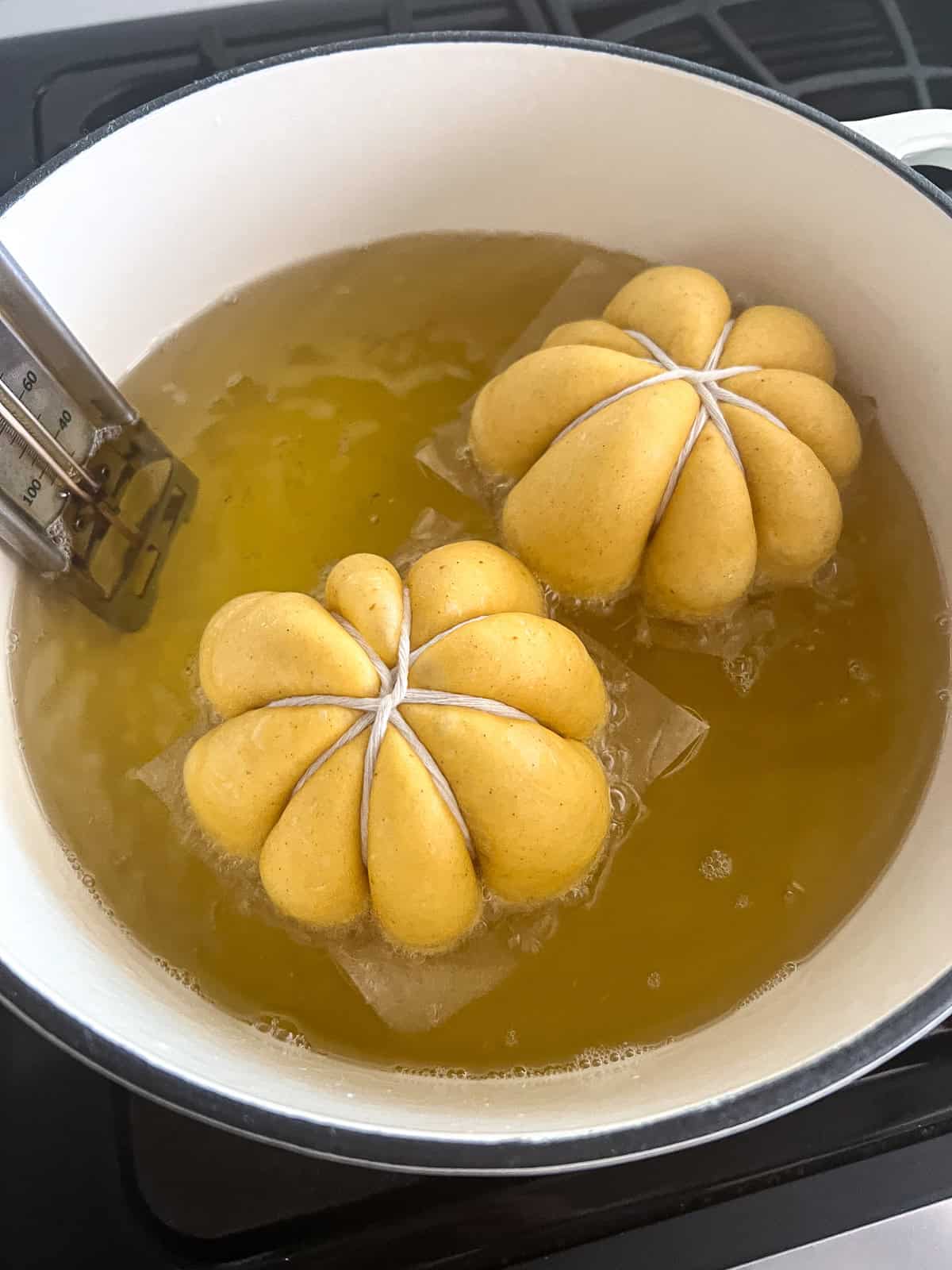 Two pumpkin donuts are fried in a large white dutch oven clipped with a candy thermometer.