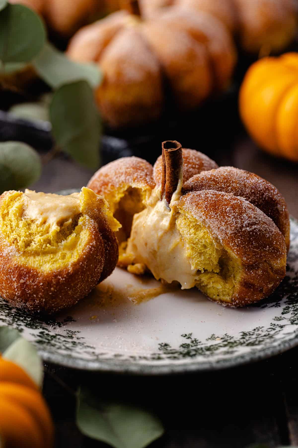 A coated and filled pumpkin shaped donut is sitting on a small dessert plate. The inside of the donut is exposed, showing the creamy pumpkin cheesecake filling and fluffy texture.