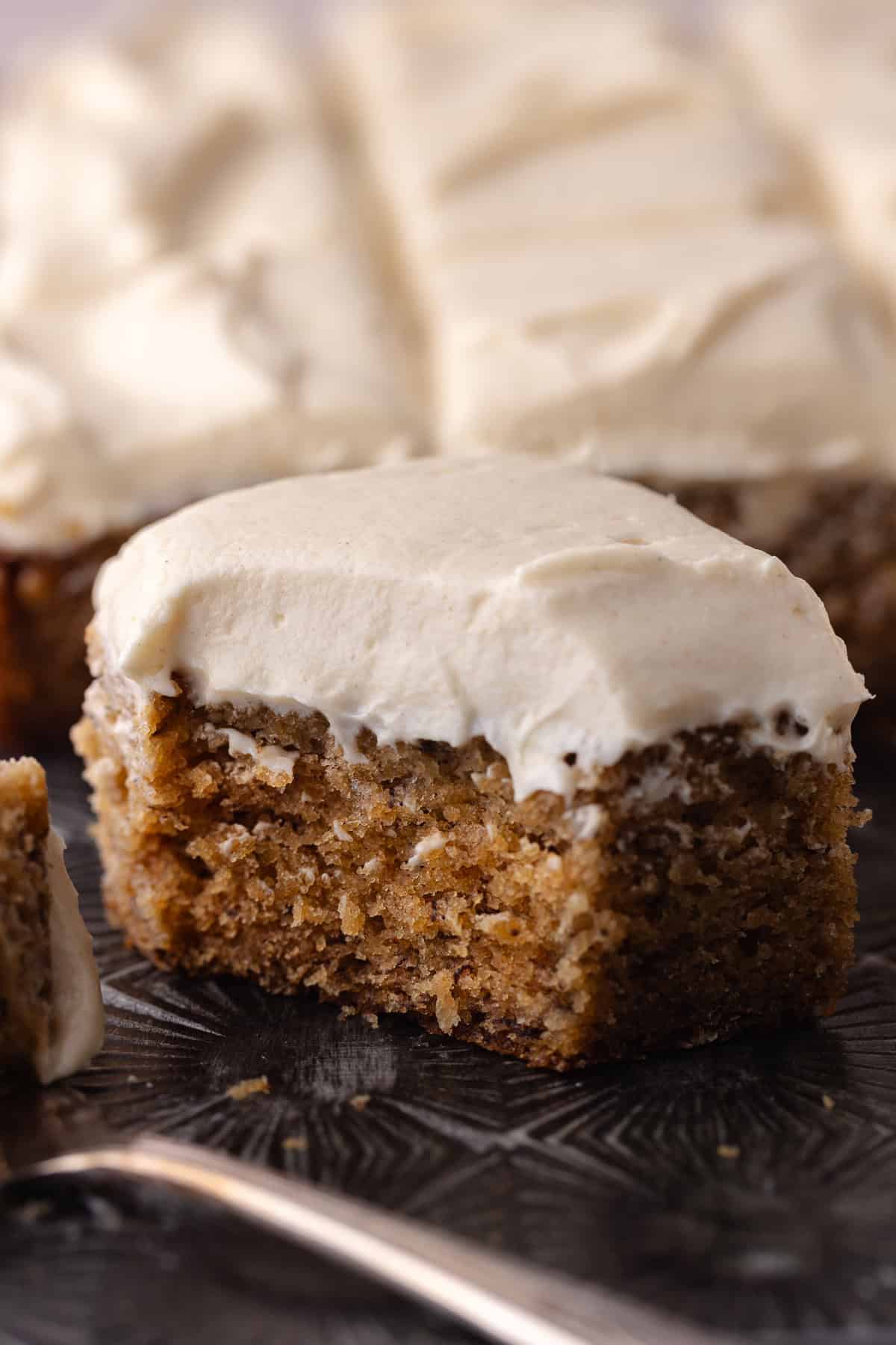 A slice of spiced banana cake with cinnamon maple cream cheese frosting sitting on a baking tray. A bite is missing.