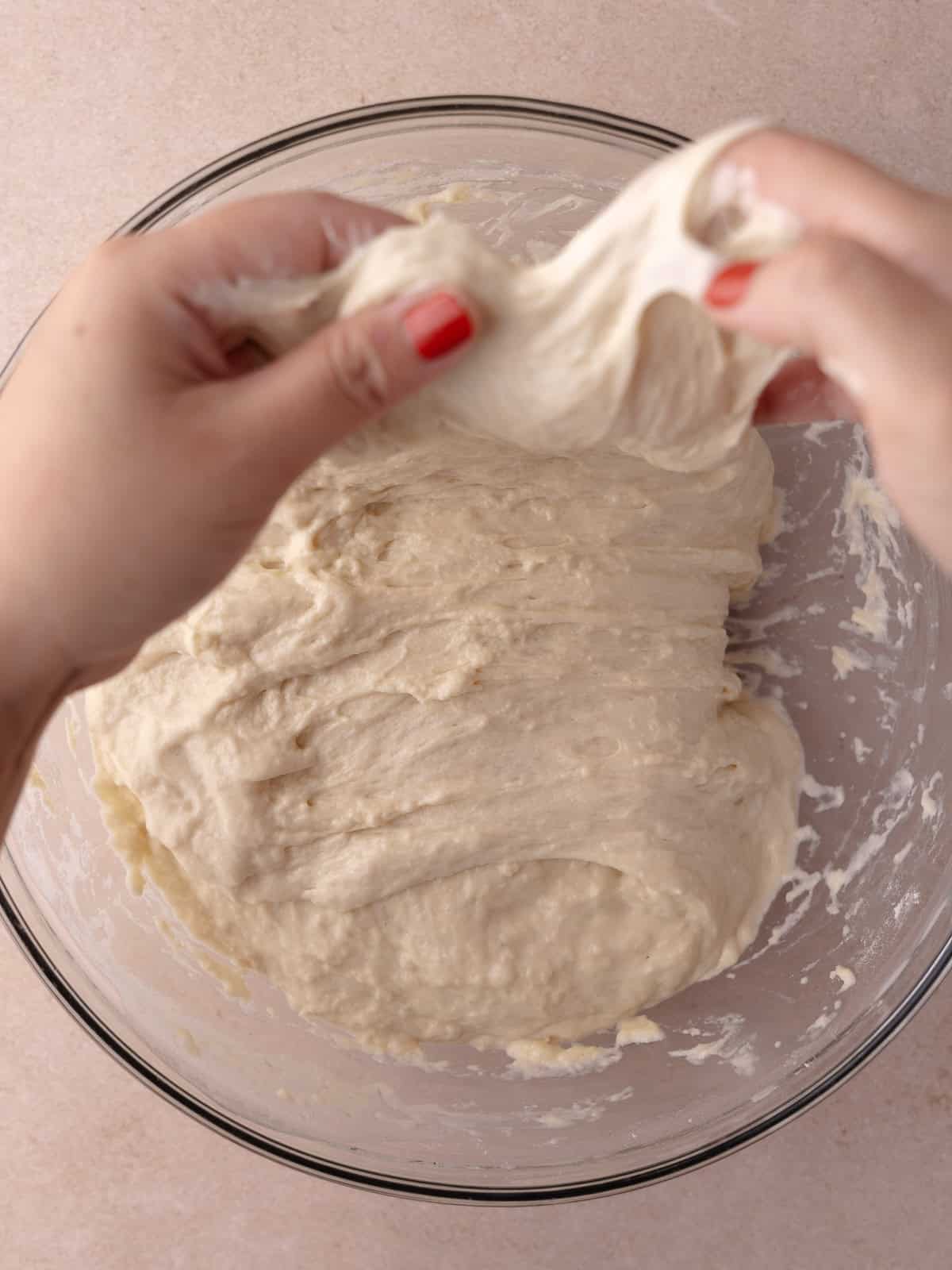 Two hands picks up a side of the dough and is stretched upwards.