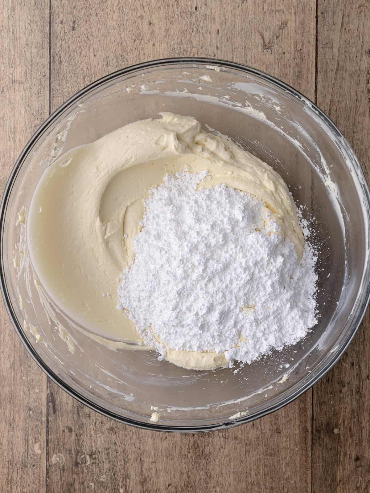 Powdered sugar is added to the butter/cream cheese mixture that is sitting in a large mixing bowl.