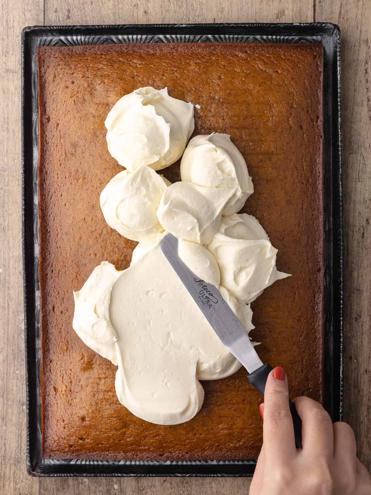 A hand spreads the maple cream cheese frosting over the top of the chai pumpkin sheet cake with an offset spatula.