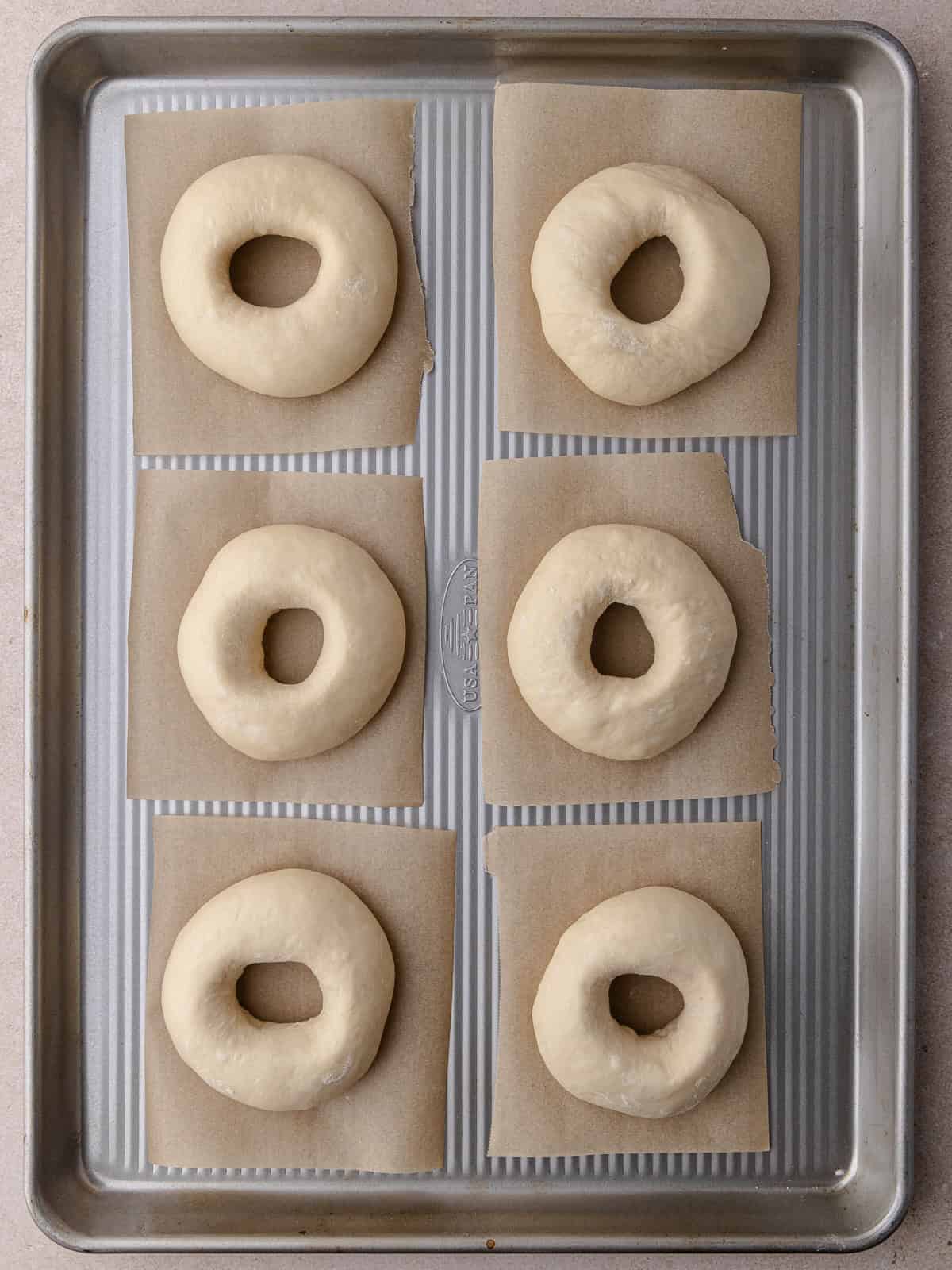 Six shaped bagels are sitting on a baking tray and are ready to proof.