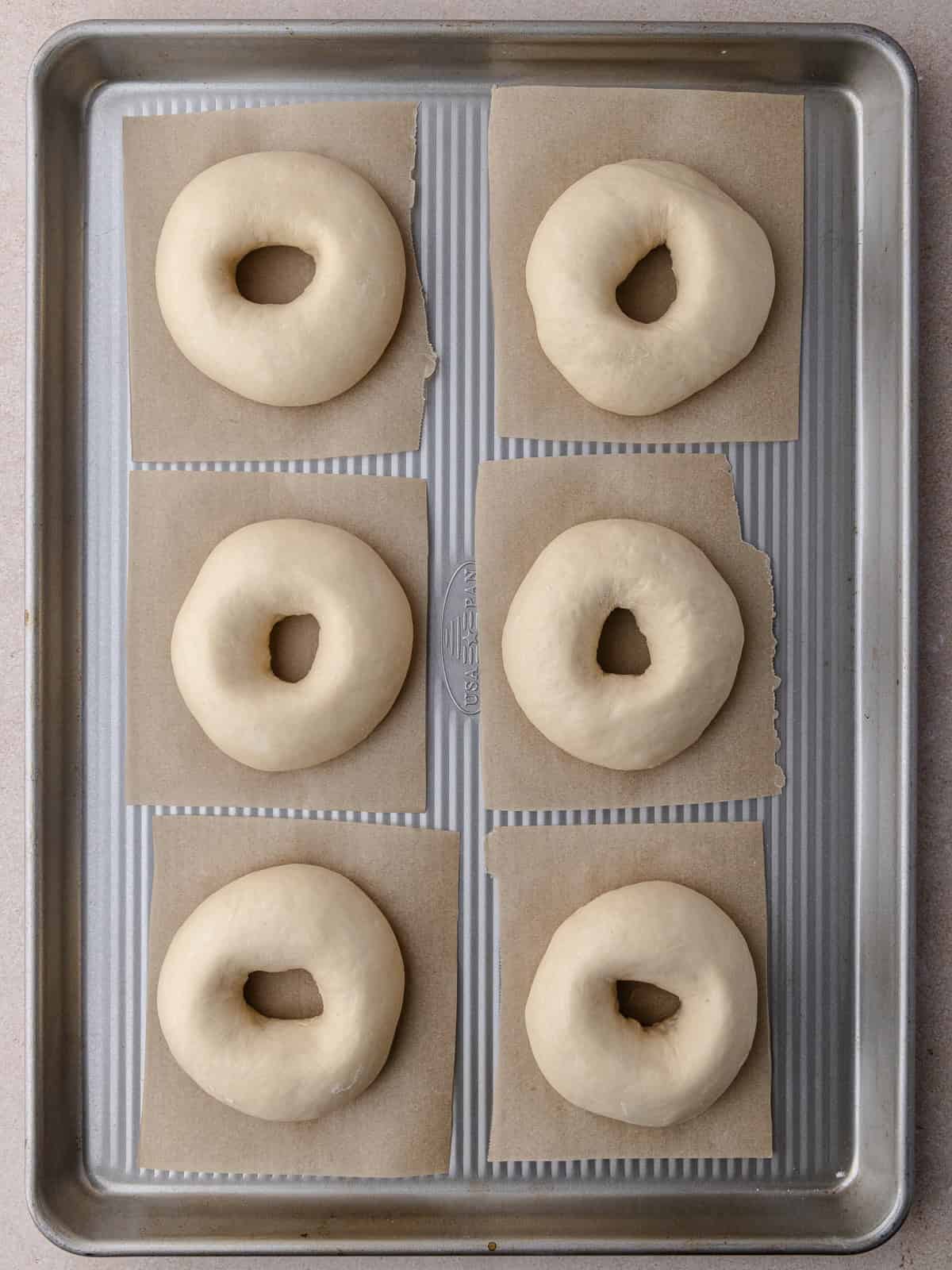 Six shaped bagels are sitting on a baking tray and have been proofed.