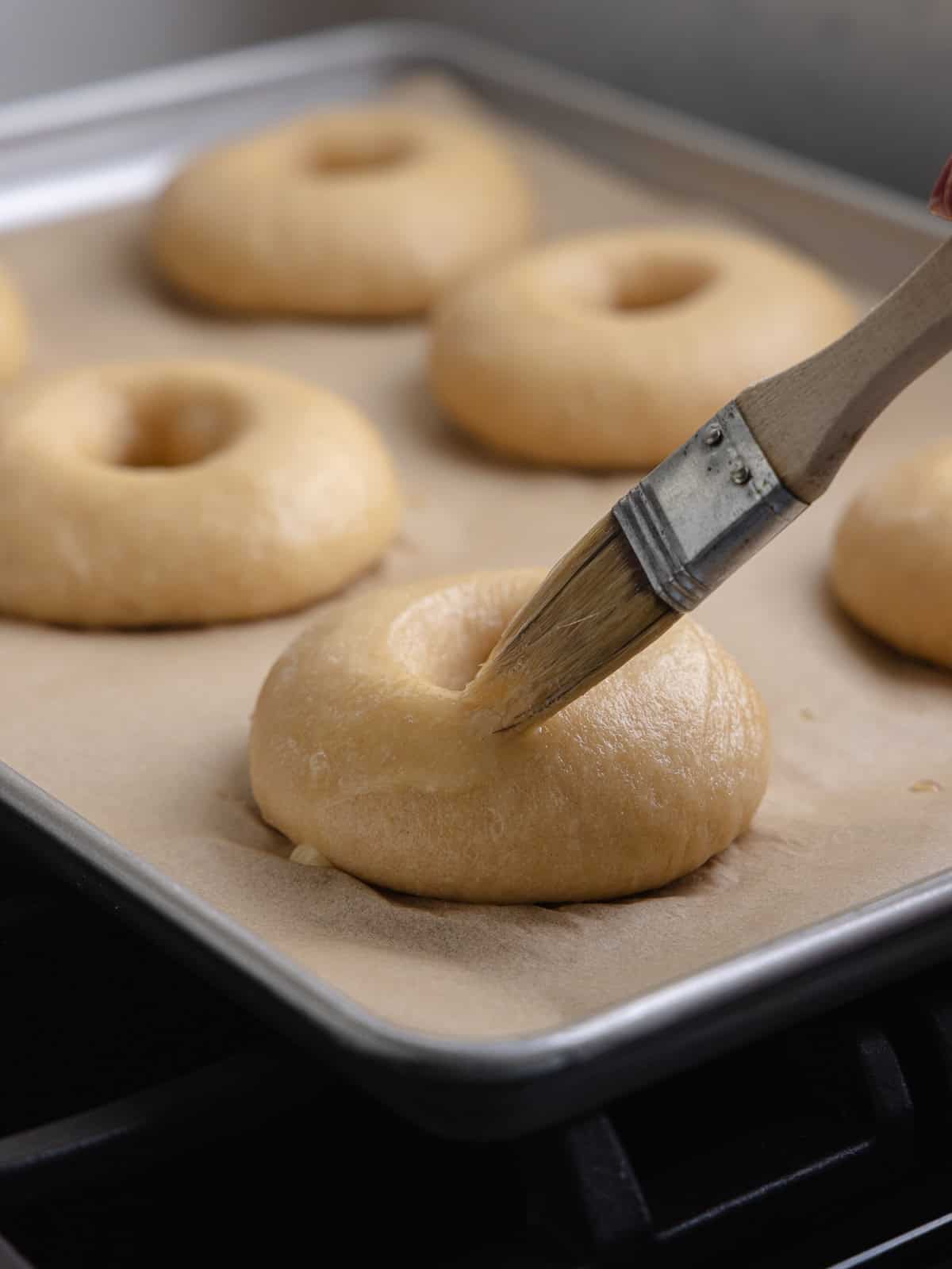 Pastry brush is painting an egg wash over the plain overnight bagels.