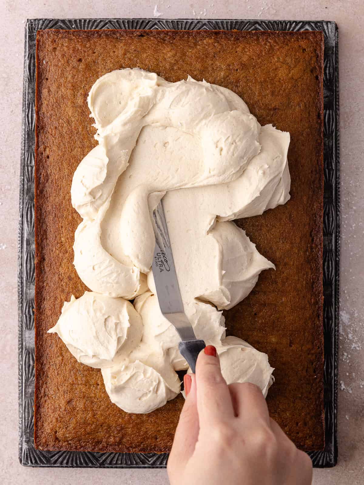 Maple cream cheese frosting is slathered on top of the spice banana cake. The sheet cake is golden brown and sitting on a baking sheet.