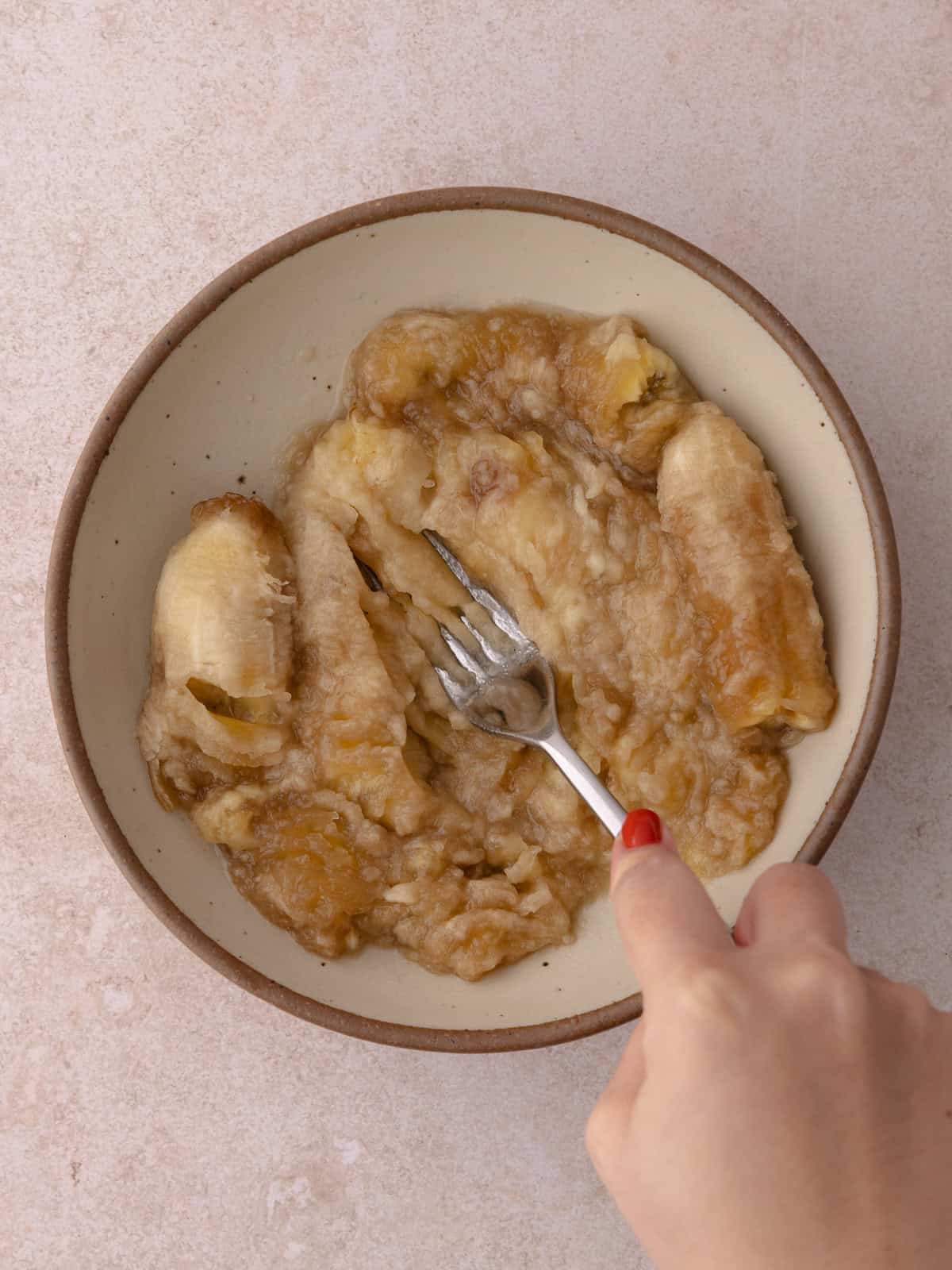 Bananas are mashed with a fork in a medium bowl.