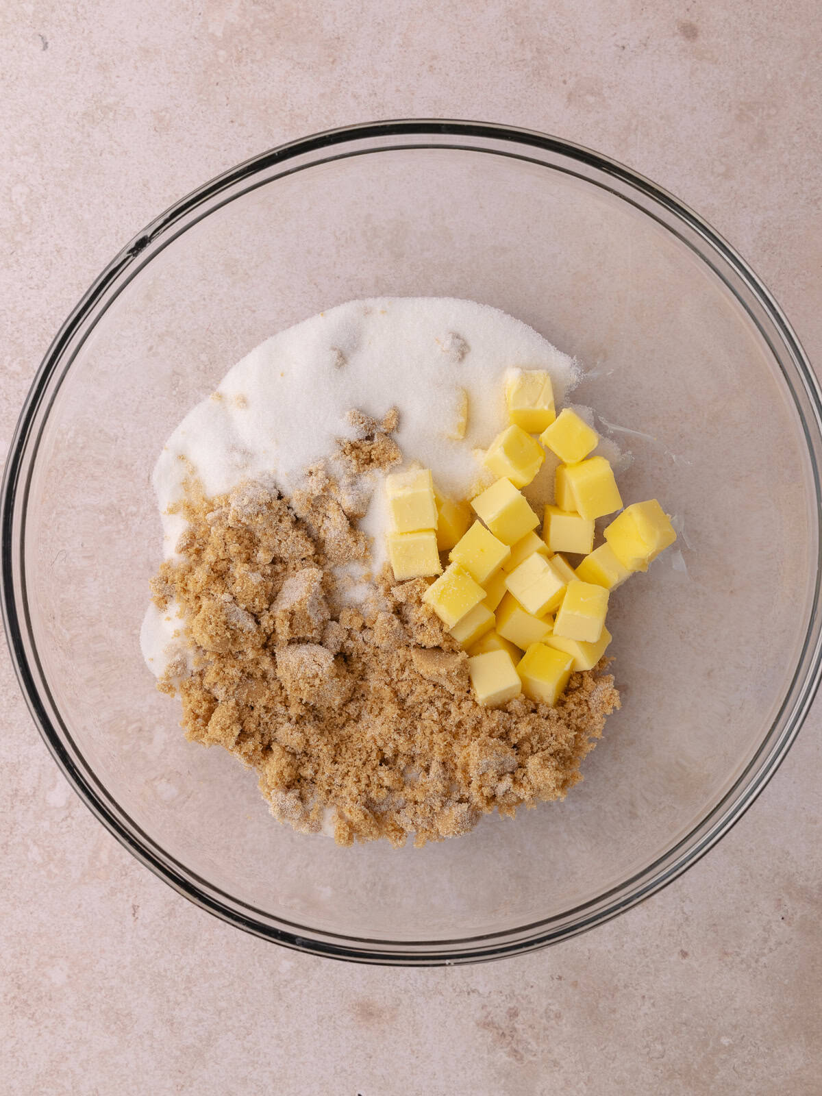 Softened cubed butter, brown sugar and white sugar are in a large glass mixing bowl.