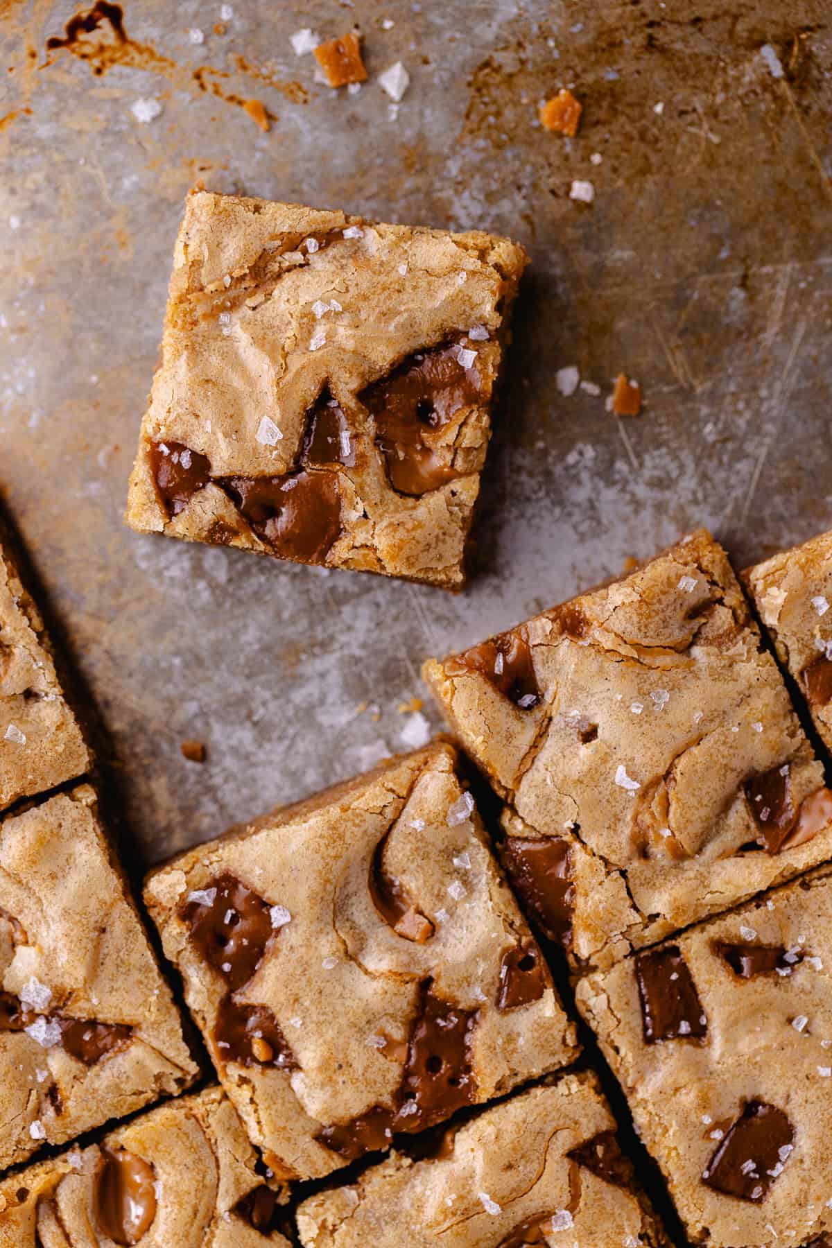 Toffee cookie butter blondies topped with flaky sea salt that are cut into squares are on a baking tray.