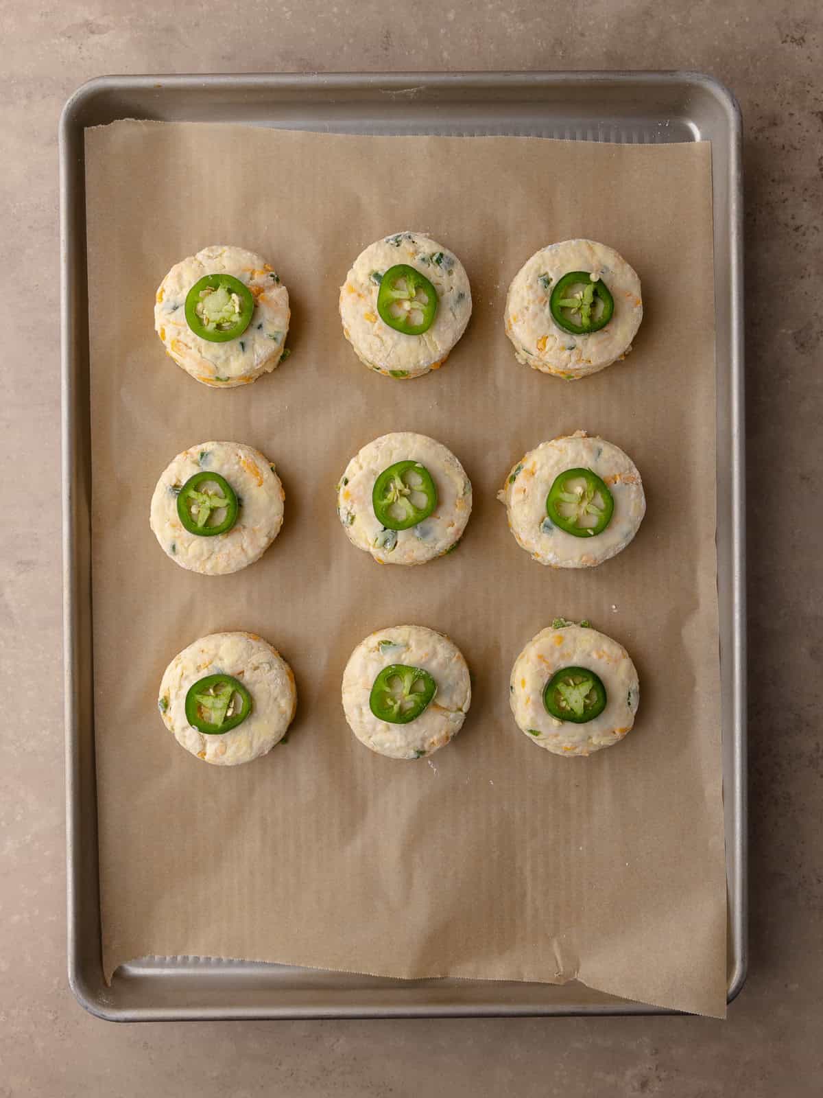 Cut out Buttermilk Jalapeño cheddar biscuits sitting on a baking tray lined with parchment paper. Biscuits are brushed with buttermilk and topped with jalapeño rings.