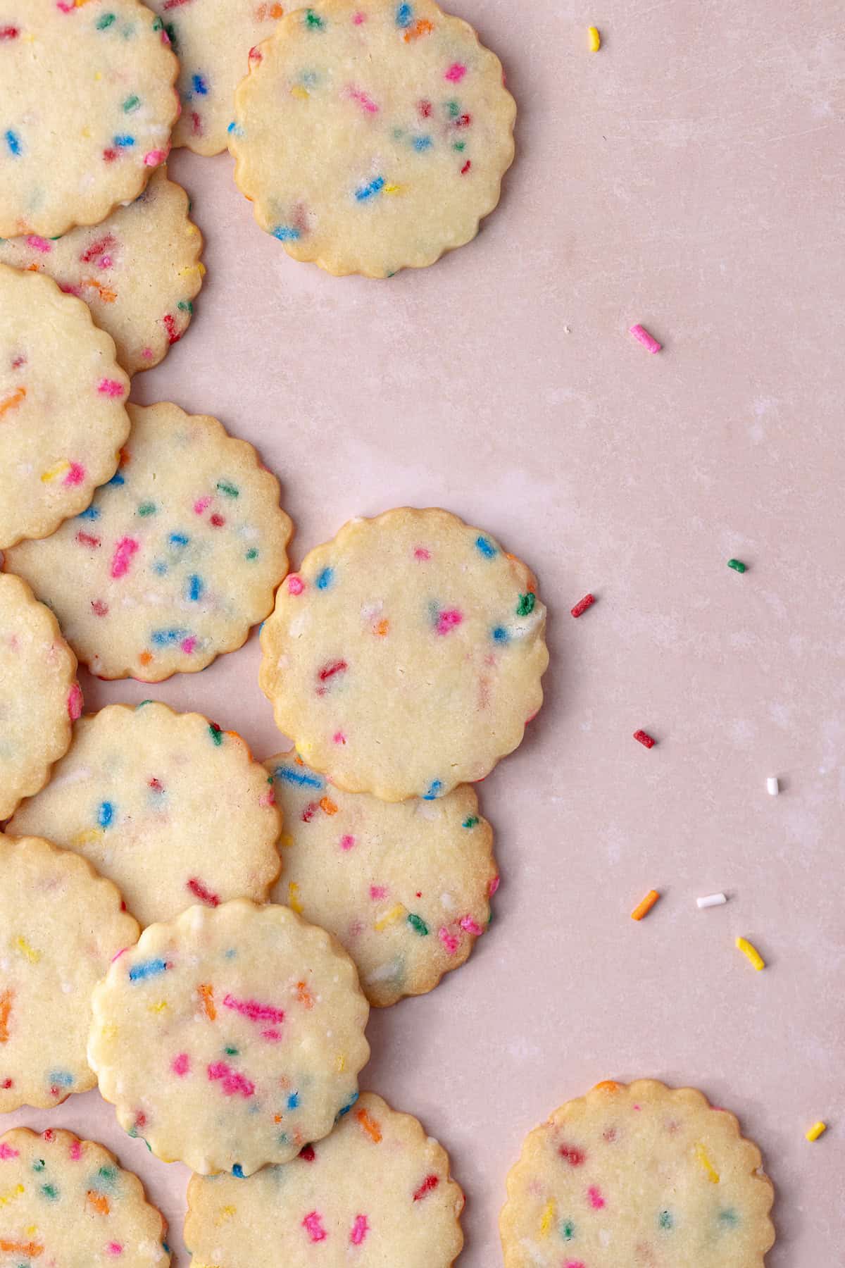 Funfetti shortbread cookies are scattered on a pink background with rainbow sprinkles.