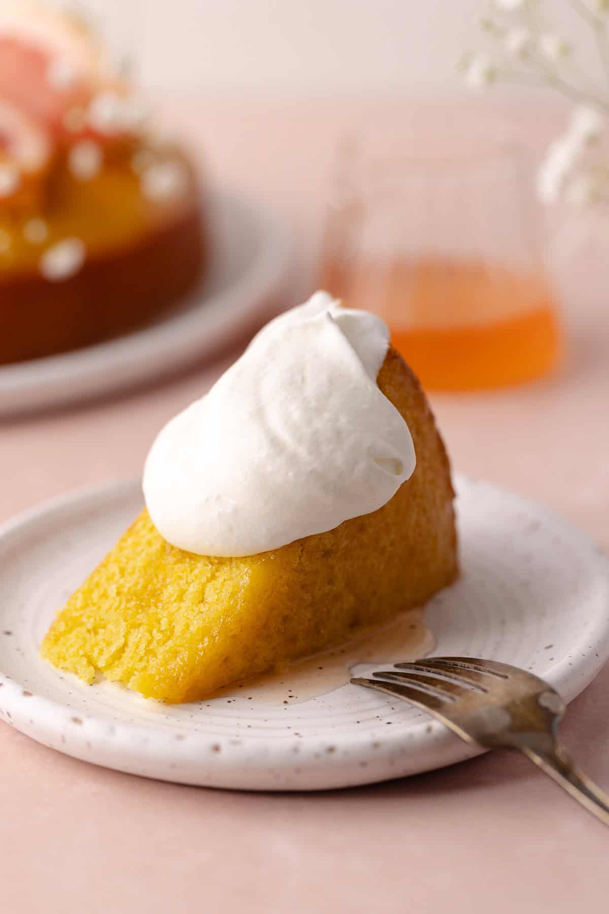 A slice of soft and moist polenta grapefruit olive oil cake sitting on a dessert plate and topped with whipped cream.