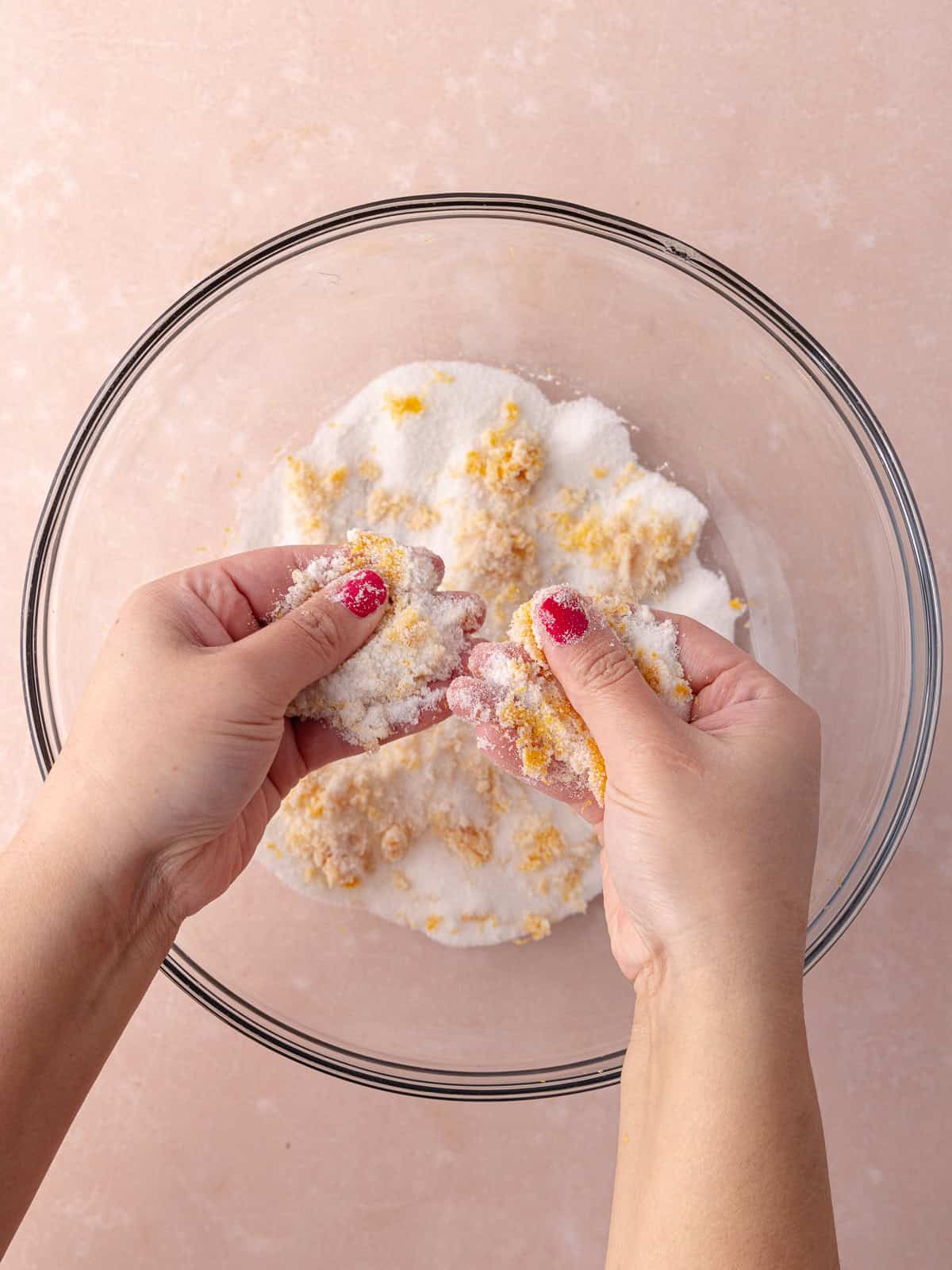 Hands are rubbing white sugar and grapefruit zest together in a large mixing bowl.