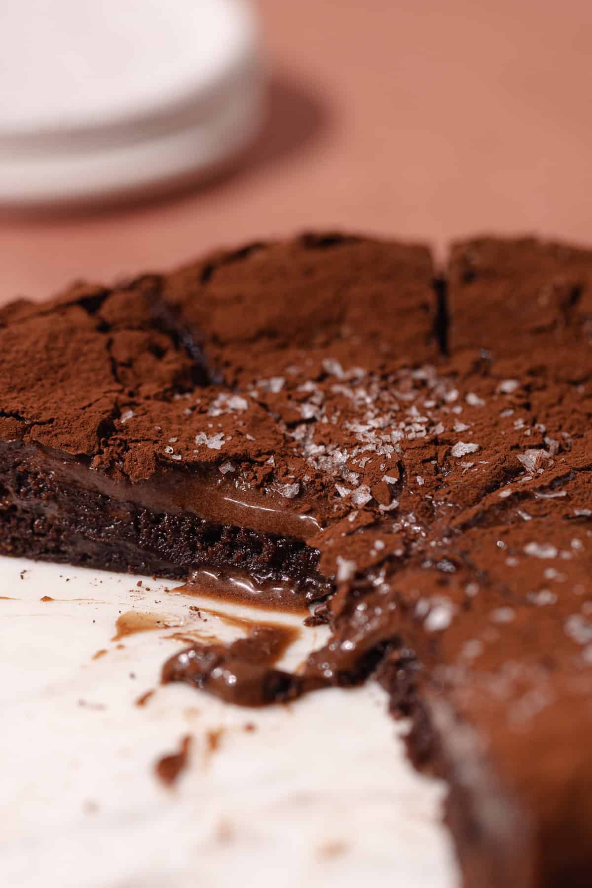 Chocolate gooey butter cake dusted with cocoa powder and topped with flaky sea salt, are cut in slices and sitting on a cake plate.