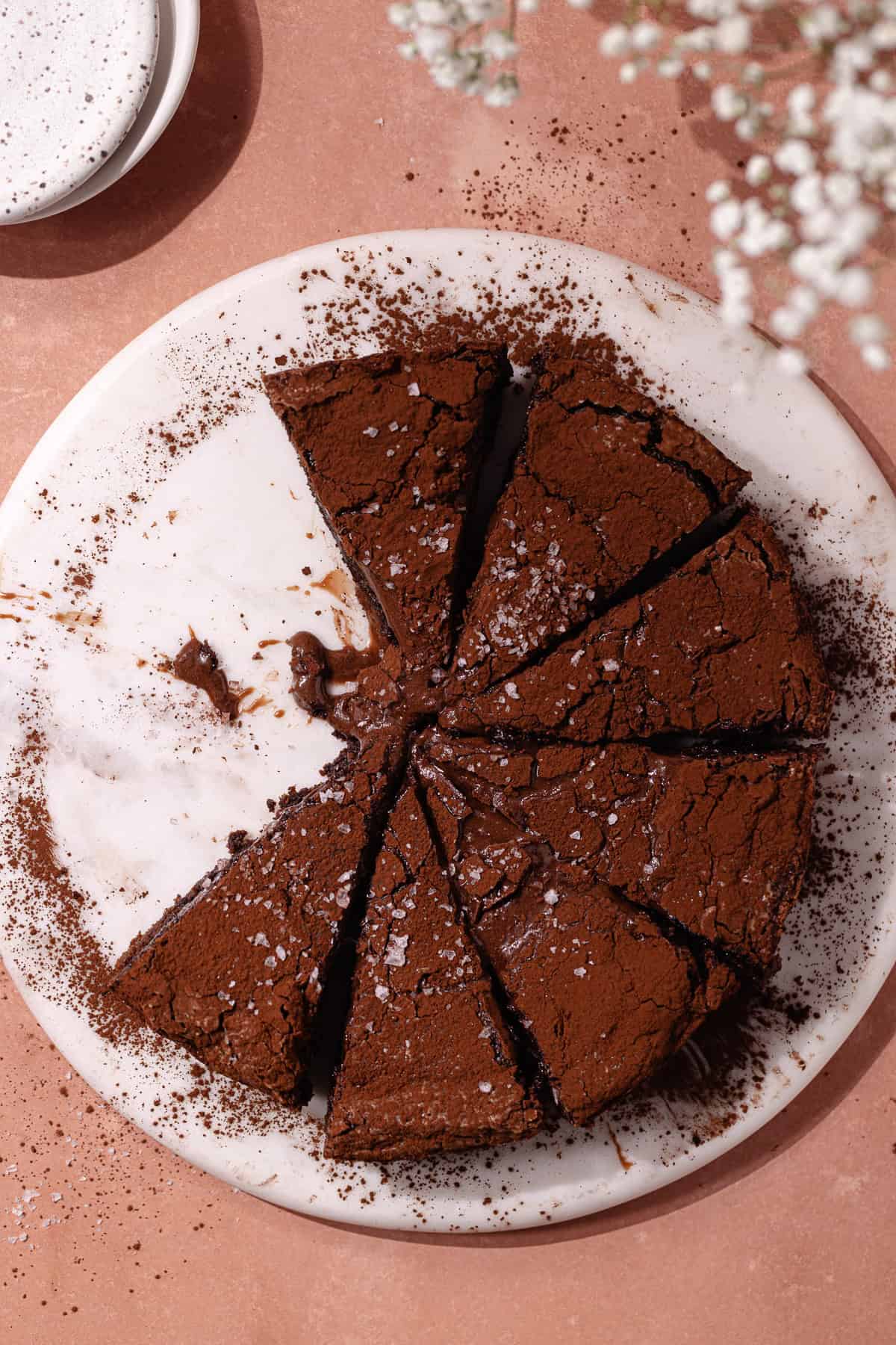 Chocolate gooey butter cake dusted with cocoa powder and topped with flaky sea salt, are cut in slices and sitting on a cake plate.