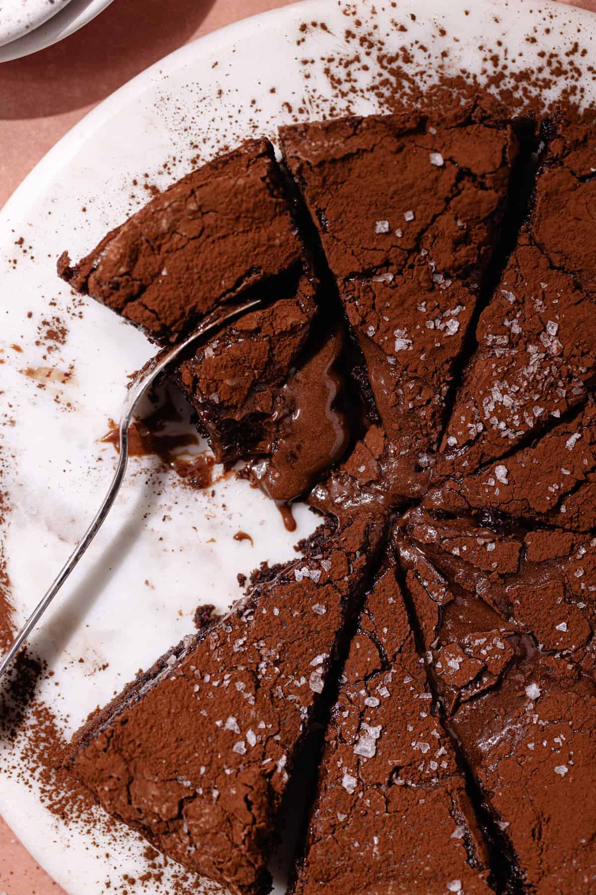 Chocolate gooey butter cake dusted with cocoa powder and topped with flaky sea salt, are cut in slices and sitting on a cake plate.