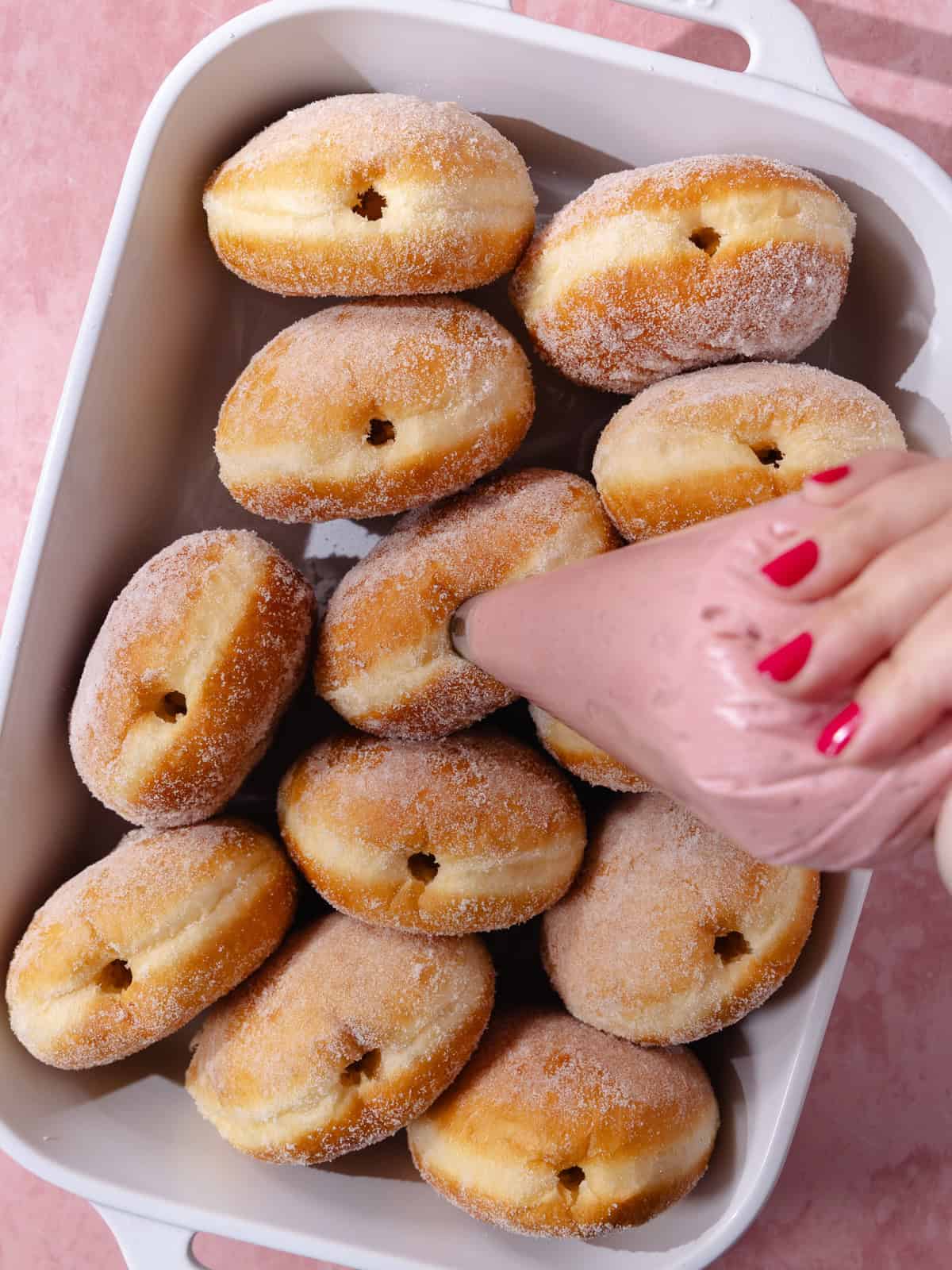 A piping bag filled with raspberry cheesecake filling is piped into donuts.