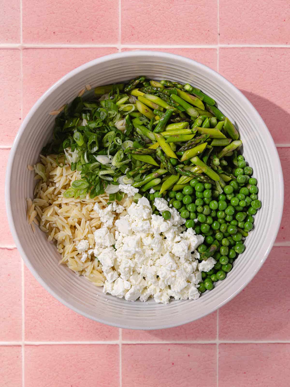 Cooked orzo, peas, asparagus, crumbled feta and chopped green onions are in a large mixing bowl.